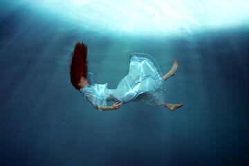 Weightlessness. Young girl with red hair, in dress gracefully falling down the ocean, showing...