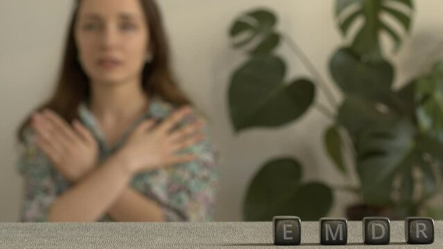 Letters EMDR written on black stone cubes. A female is touching and tapping her shoulders in a blurred background. Eye Movement Desensitization and Reprocessing Psychotherapy Treatment concept.