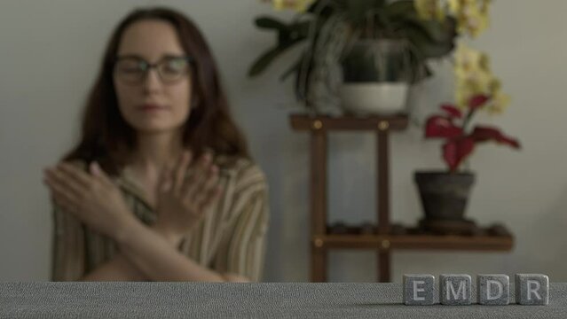 Letters EMDR written on gray stone cubes. A woman with glases is touching and tapping her shoulders in a blurred background. Eye Movement Desensitization and Reprocessing Psychotherapy Treatment conce