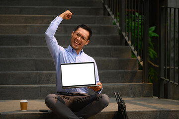 Cheerful businessman holding laptop with blank screen and raising hand in yes gesture