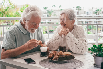 Cheerful bonding senior retired couple enjoy breakfast together sitting outdoors on home terrace....