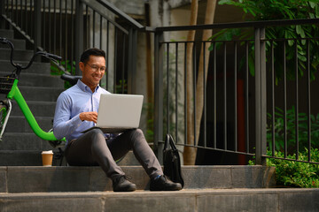 Positive millennial male entrepreneur sitting on the stairs in the city and using laptop