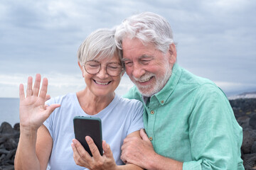 Video call concept. Happy senior couple sitting outdoors by the sea waving hand in video connection...