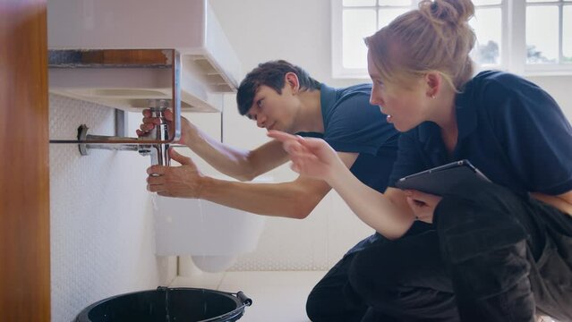 Female plumber holding digital tablet with male trainee fixing leaking pipe in bathroom sink - shot in slow motion