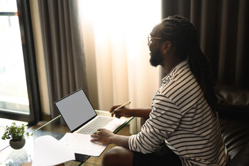 Side view of African American man writing notes working remotely on laptop at home