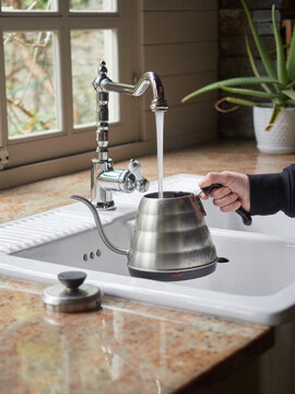 hand holding a kettle handle refilling water coming out of stainless steel kitchen faucet. Tea preparation.