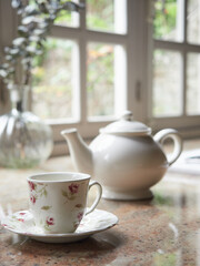 Close-up of porcelain tea cup and teapot on kitchen countertop indoors in a cozy cottage. Window out of focus in the background.