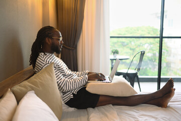 Young African American man using laptop computer on bed , working at his hotel room