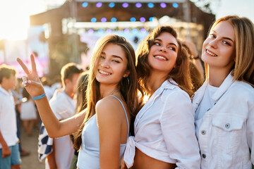 Group of happy girls at summer beach music fest, smiling, dancing in sun, flashing peace sign. Friends enjoy live concert, sunset light in background. Casual trendy outfits, fun, youth culture vibe.