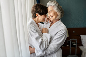 Senior lesbian couple embrace by window, sharing tender moment.
