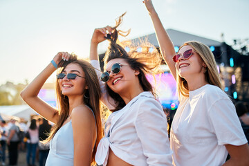 Girls in sunglasses enjoy music at sunny beach fest. Friends dance, laugh with stage behind. Casual...