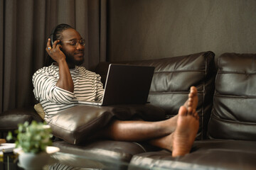 Relaxed African man in headphones using laptop on couch and listening music with closed eyes
