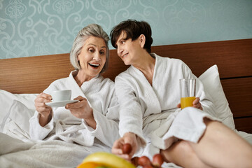 Two women in white robes, a senior lesbian couple, sit peacefully on a bed.