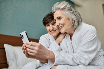 Senior lesbian couple share a tender moment while viewing a cell phone on a bed. - Powered by Adobe