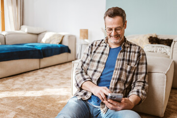 Happy Middle-Aged Man Sitting on Floor, Laughing While Typing on Smartphone