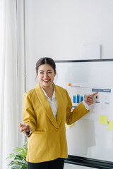 Confident Businesswoman Presenting: A young professional woman in a yellow blazer stands confidently in front of a whiteboard, presenting data with a warm smile and an engaging demeanor.