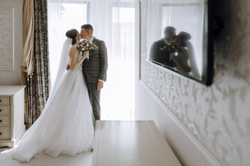 A bride and groom are kissing in front of a mirror. The bride is holding a bouquet of flowers