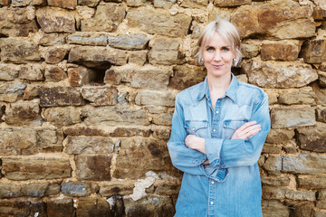 Confident young woman with short blonde hair standing with crossed arms against a stone wall, exuding a relaxed and friendly vibe