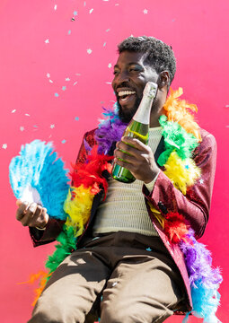 Cheerful non-binary person holding champagne bottle and hand fan against pink background