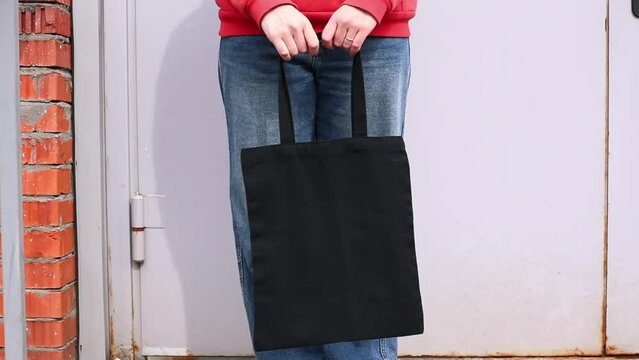 Woman in red hoodie holding tote canvas blank eco bag on street brick wall background. Female consumer hold black textile shopper. template or place for your design, logo, text