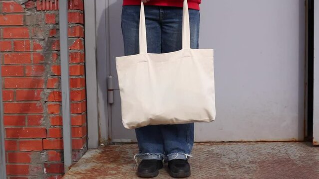 Woman in red hoodie holding tote canvas blank eco bag on street brick wall background. Female consumer hold white textile shopper. template or place for your design, logo, text