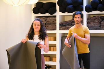Two people are holding a yoga mat in a room with shelves full of yoga material. Scene is calm and focused, as the people are likely preparing for a yoga session.