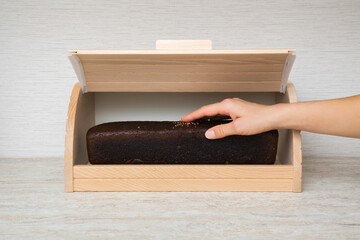 Young adult woman hand holding fresh dark brown rye bread and taking out from wooden bread box on table top at home kitchen. Closeup. Front view. - 800102287
