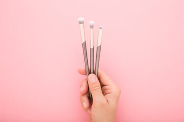 Young adult woman hand fingers holding and showing three new different makeup brushes with soft bristles on light pink table background. Pastel color. Female beauty product. Closeup. Top down view. - 800102405