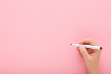 Young adult woman hand holding black white color pen and writing on light pink table background. Pastel color. Closeup. Empty place for text. Top down view.