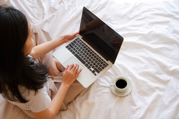 top view of Asian Woman sitting on the bed with cup of coffee and working on her laptop.