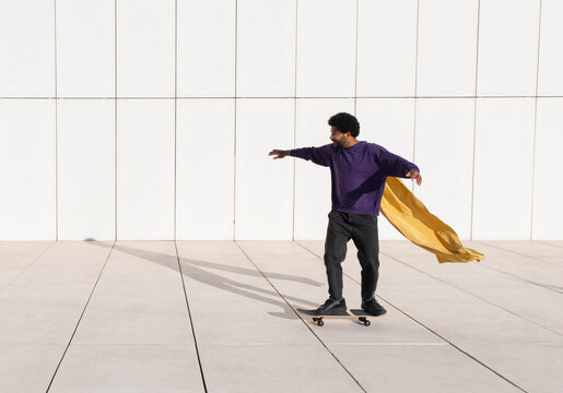 Young man skateboarding in front of white wall wearing a cape