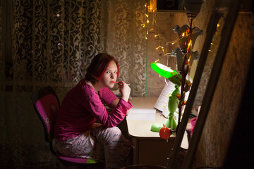 A schoolgirl girl with red hair sits at a table with a table lamp and a luminous garland, reads a...