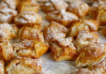 Freshly baked pastries, golden brown in color on a baking sheet. They are generously sprinkled with powdered sugar giving them a sweet appearance. The texture of the pastries looks flaky and crispy.