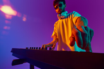 Obraz premium Young man, lost in the rhythm, mixes tracks on a DJ mixer, fueling energy of crowd against gradient pink purple background in neon light. Concept of music, performance, festival, live concert