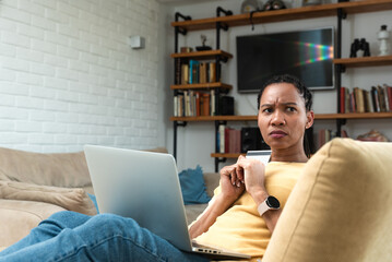 Confused woman sitting on couch holds credit card use laptop looking at device screen having debt...
