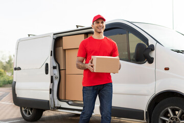 Positive young delivery man in red uniform holding cardboard box beside parked delivery van,...
