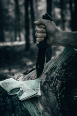 Man hand in forest holds hunting knife against background terrain map and compass. Concept survival...