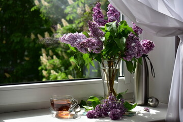 a bouquet of lilac flowers and tea from a thermos in a glass cup on the windowsill