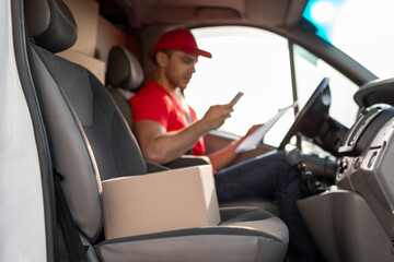 Courier man checking customer address using cellphone, sitting at drivers seat in van, focus on parcel nearby