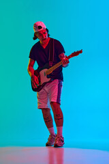 Young man, musician emotionally playing electric guitar with intensity and focus against blue, cyan background in neon light. Concept of music, performance, festival, live concert