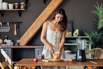 Charming curly-haired brunette girl in linen sundress prepares fruit smoothie in blender in kitchen.