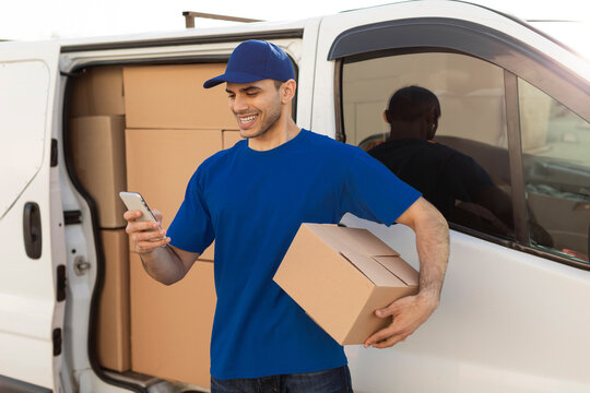 Happy young delivery man holding parcel box and using cellphone while standing near van outdoors