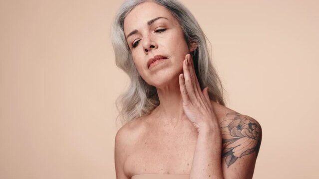 Graceful senior woman with a tattoo touches her skin, symbolizing natural beauty and skincare in a studio setting.