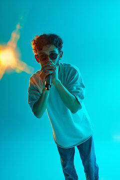 Rap, beatbox music. Young man in white t-shirt, sunglasses performing with microphone against blue, cyan background in neon light. Concept of music, performance, festival, live concert