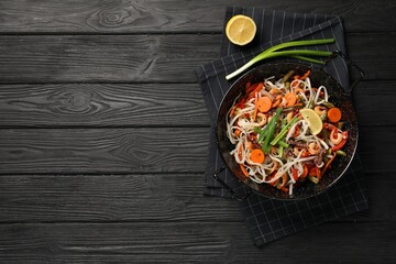 Shrimp stir fry with noodles and vegetables in wok on black wooden table, top view. Space for text
