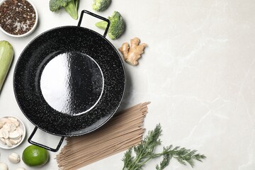 Empty iron wok surrounded by raw ingredients on light table, flat lay. Space for text