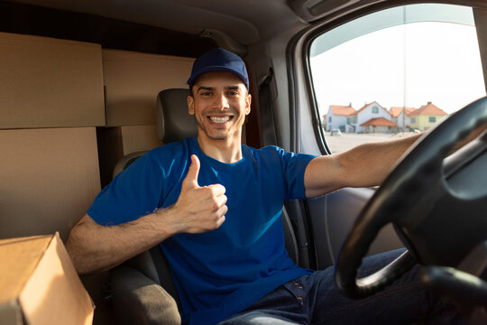 Parcel shipping. Smiling young deliveryman driver gesturing thumbs up, sitting in car with cardboard boxes and packages