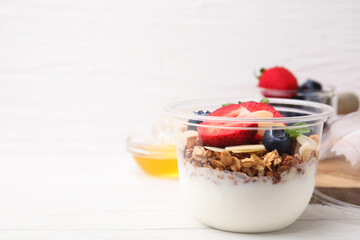 Tasty granola with berries, yogurt and almond flakes in plastic cup on white wooden table. Space for text