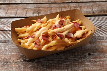 Tasty potato fries, cheese sauce and bacon in paper container on wooden table, closeup