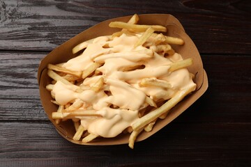 Tasty potato fries and cheese sauce in paper container on wooden table, top view